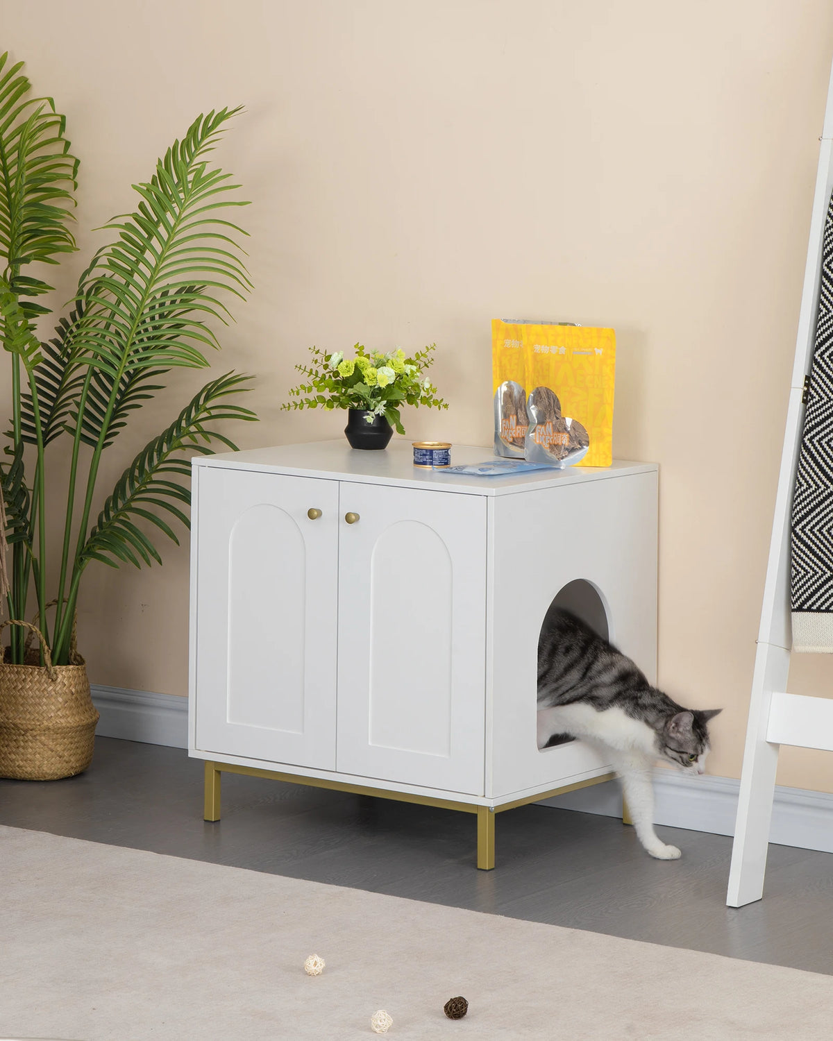 Meuble cache-litière blanc à pieds dorés, un chat sort par l'entrée latérale